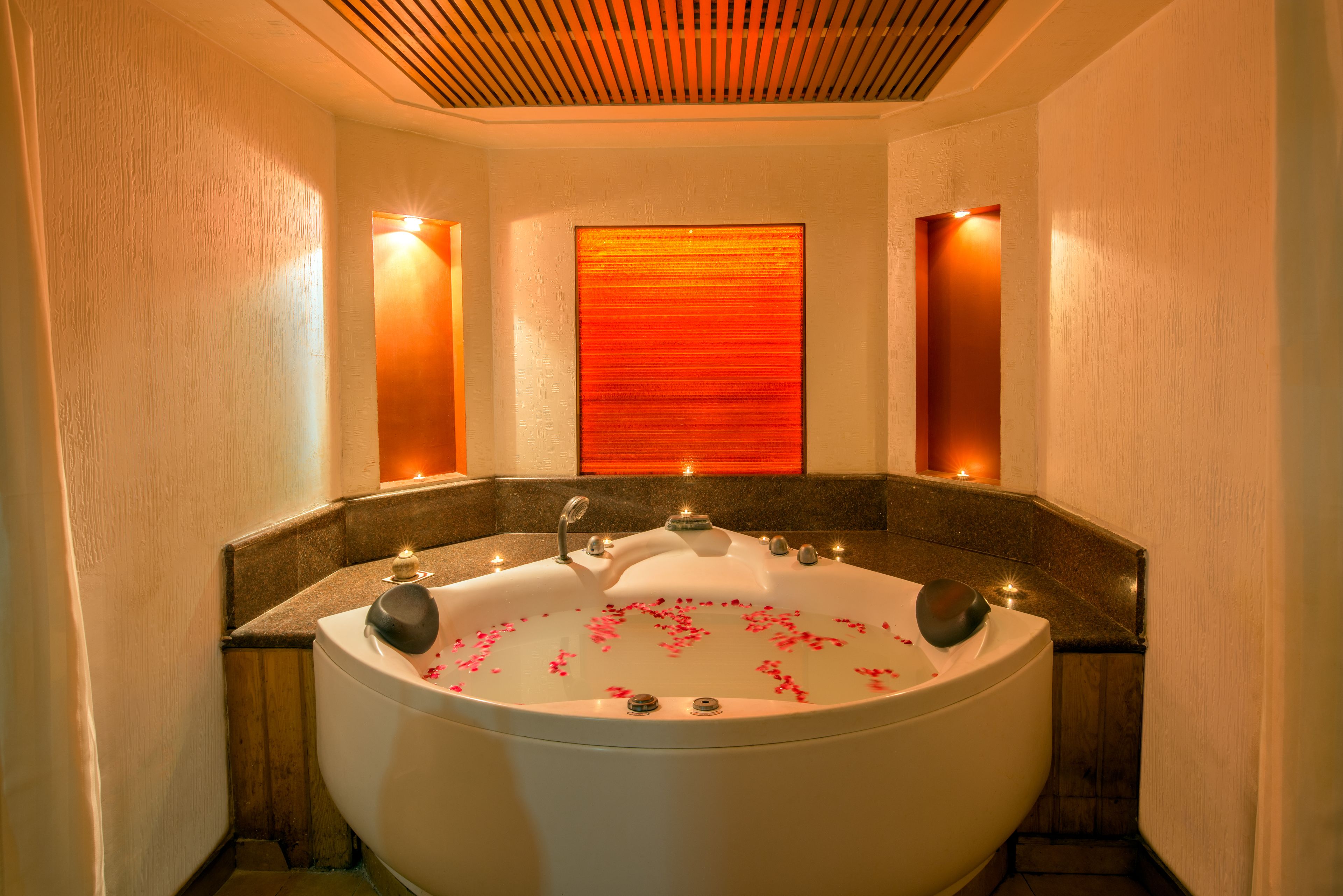 A large freestanding bathtub with rose petals in the water, surrounded by a wooden bench and a wall-mounted light fixture.
