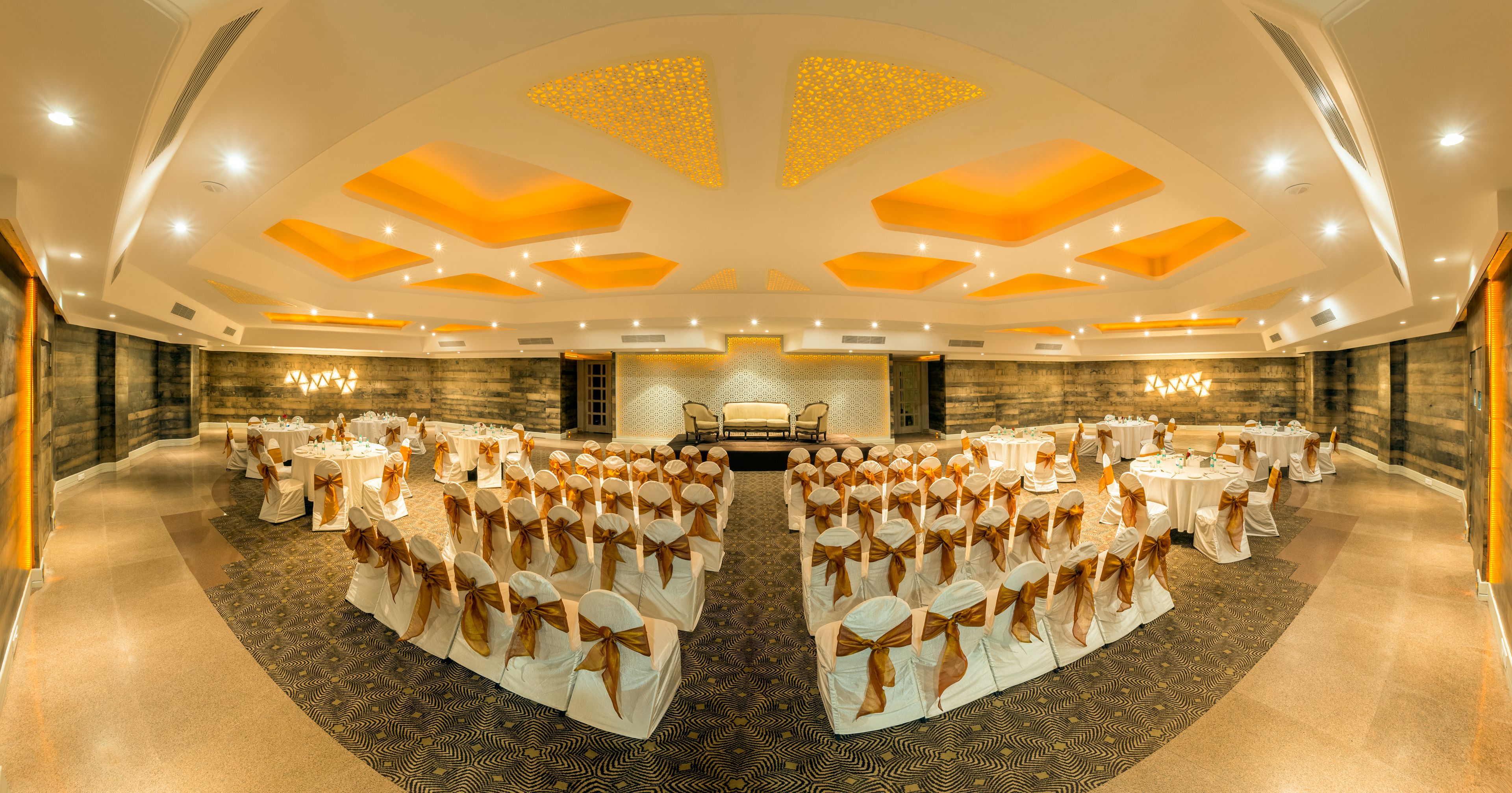 A banquet hall with rows of round tables, each with white tablecloths and gold sashes, and a central stage area.