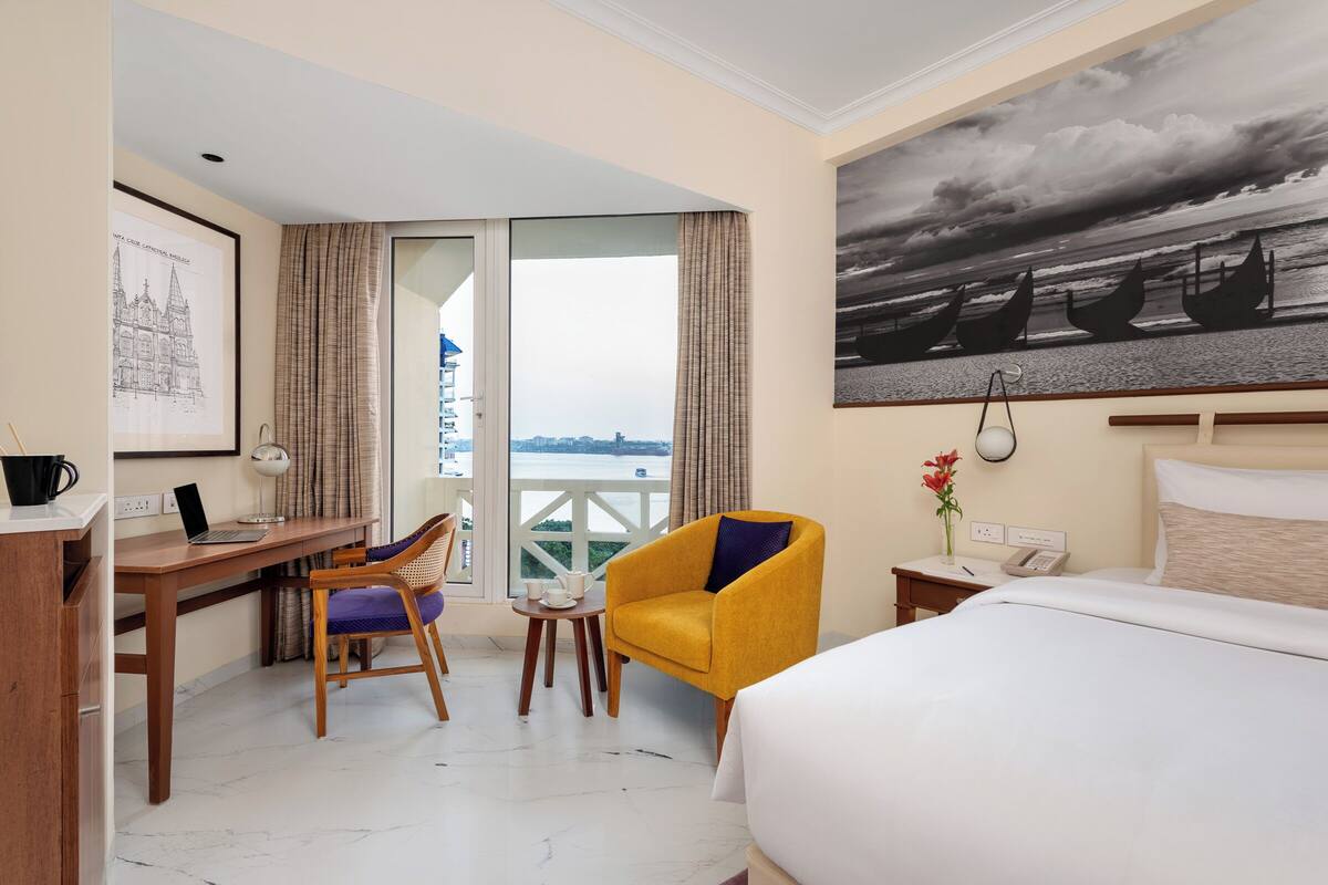 A hotel room with a bed, a desk with a laptop, a chair, a small table with a phone and a vase of flowers, and a balcony with a view of the city.