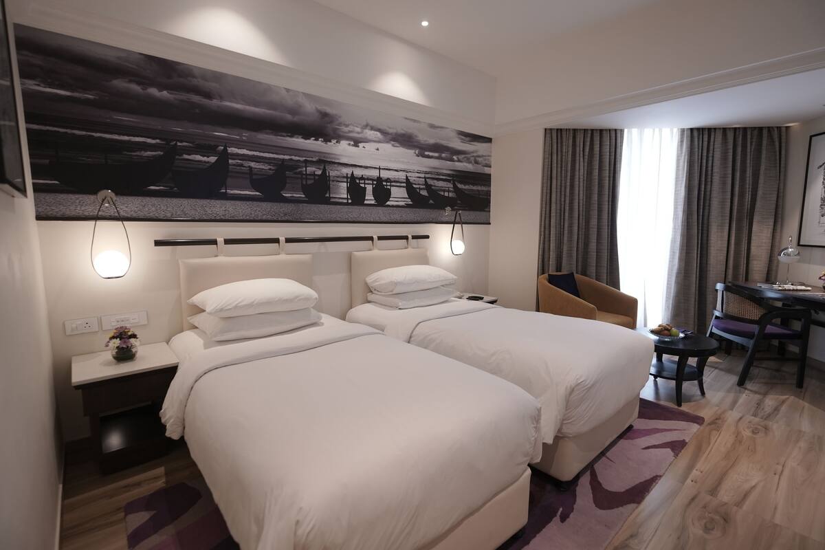 A hotel room with two beds, a desk, and a chair. There is a painting on the wall and a window with curtains.