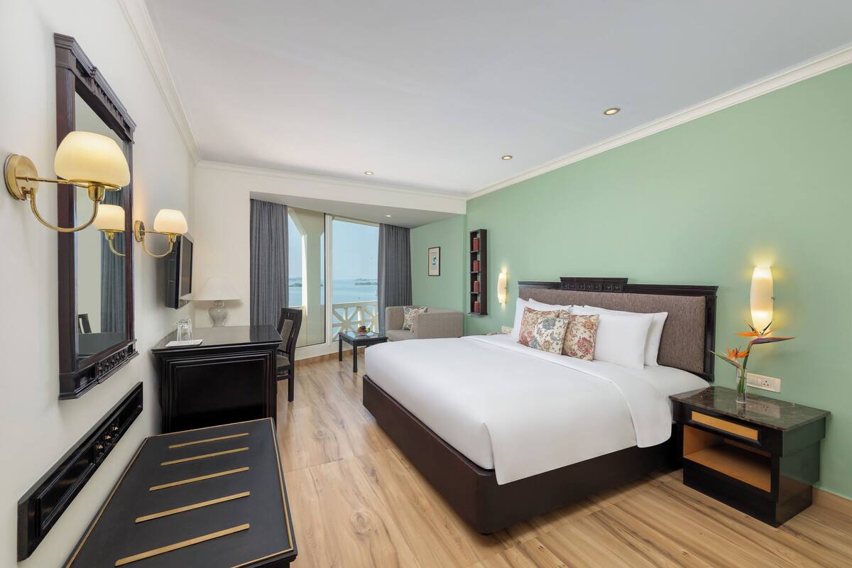 A hotel room with a large bed, bedside tables, a desk, a chair, a mirror, and a view of the sea.