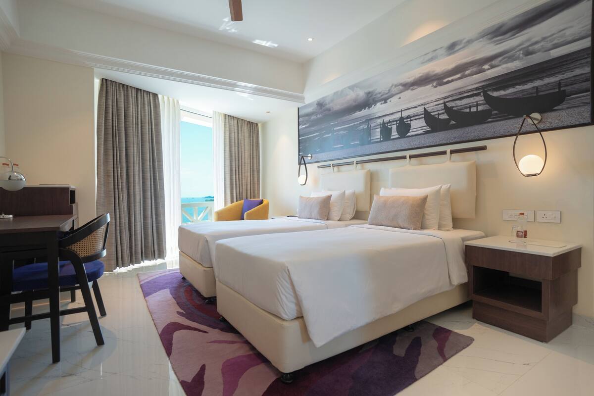 A hotel room with two beds, a desk, and a chair. There is a large window with curtains, a painting of boats on the wall, and a nightstand with a lamp.