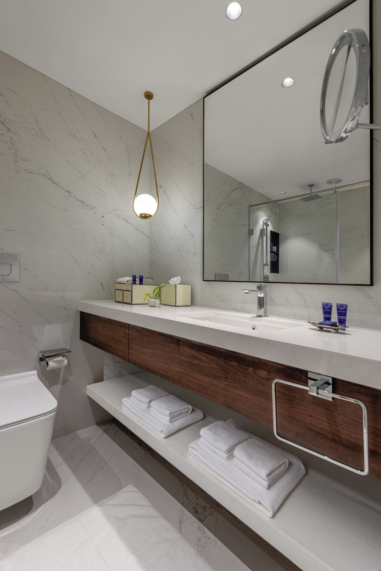 A modern bathroom with a marble wall, a white sink, a large mirror, and a wooden vanity.