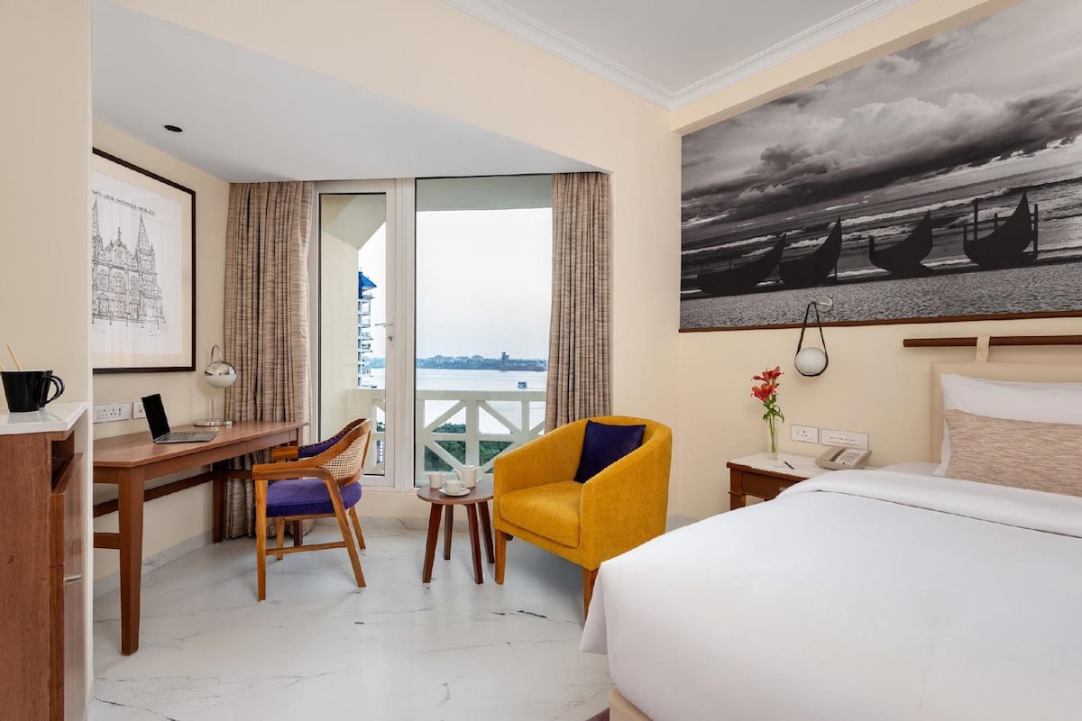 Ein Hotelzimmer mit Bett, Schreibtisch, Stuhl, einem kleinen Tisch, einer Lampe, einem Telefon, einer Vase mit Blumen, einem Gemälde und Meerblick vom Balkon aus.