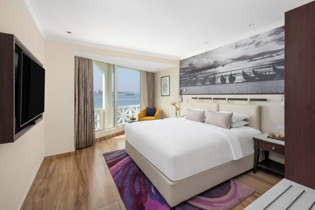 A modern hotel room with a large bed, a flat-screen TV, a balcony with a view of the sea, and a painting on the wall.