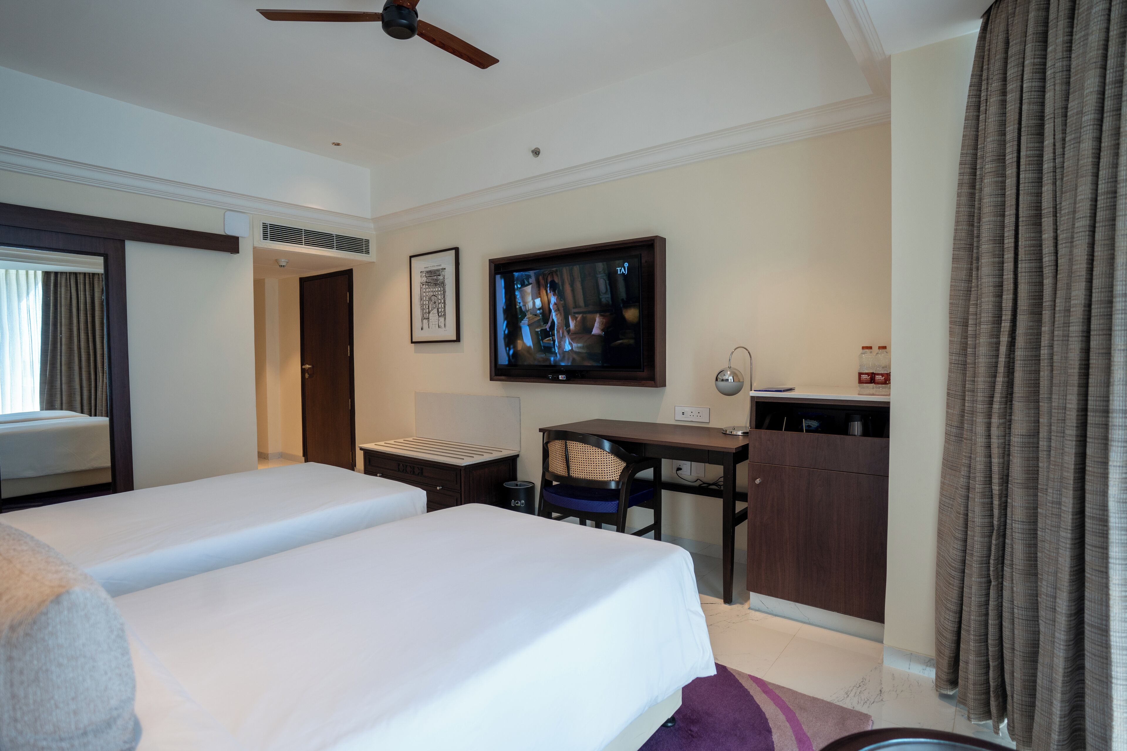Habitación de hotel con dos camas, un escritorio, televisión y ventana con cortinas.