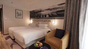 Habitación de hotel con una cama grande, un sofá, una mesita con frutero y un cuadro de un barco y un atardecer en la pared.