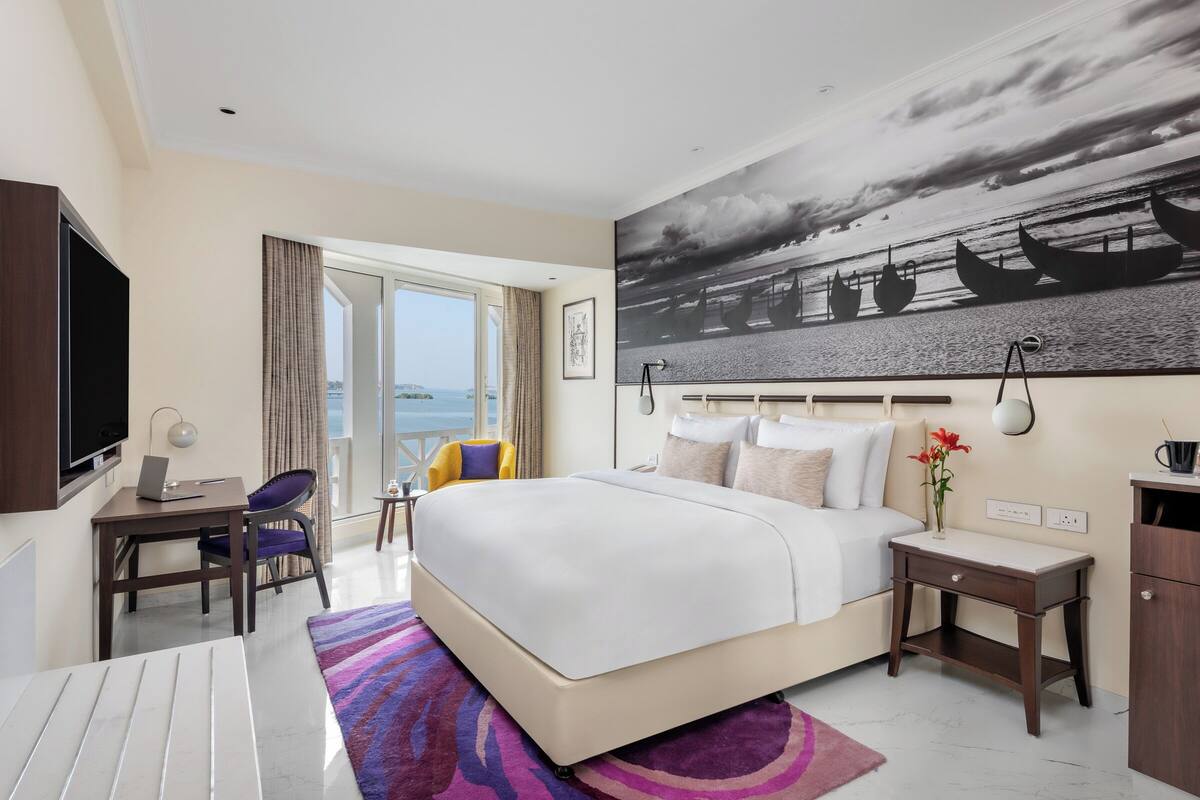 A hotel room with a large bed, a desk with a laptop, a TV, and a view of the sea.
