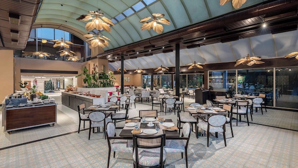 A spacious indoor dining area with multiple tables and chairs, a central bar area, and a high ceiling with natural light. - Vivanta Ernakulam, Marine Drive (Kanayannur)