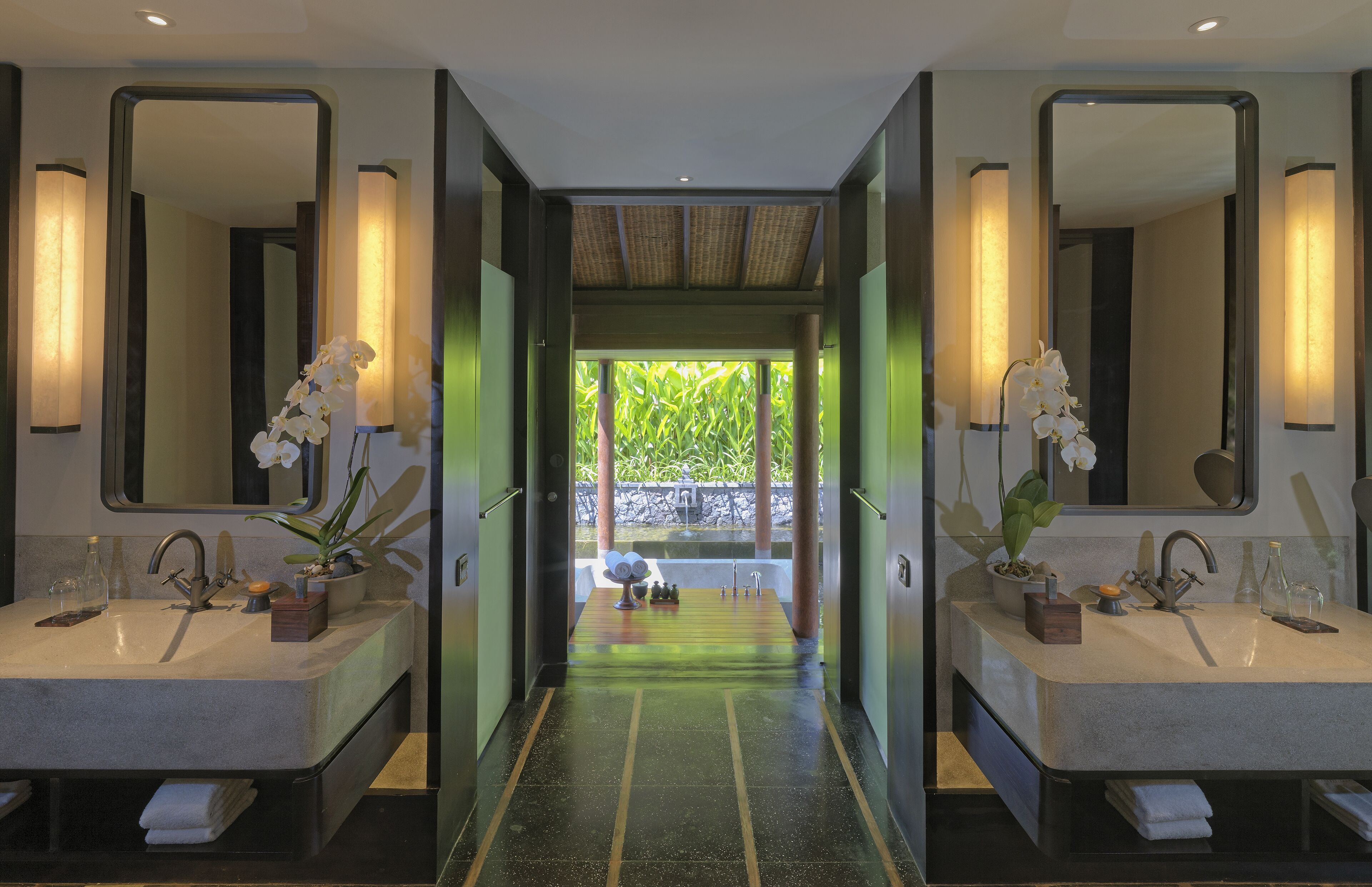 A modern bathroom with two sinks, large mirrors, and a view of greenery through a glass door.
