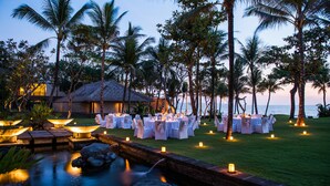 An outdoor wedding venue with tables set up for guests, surrounded by palm trees and a small pond with lighting.