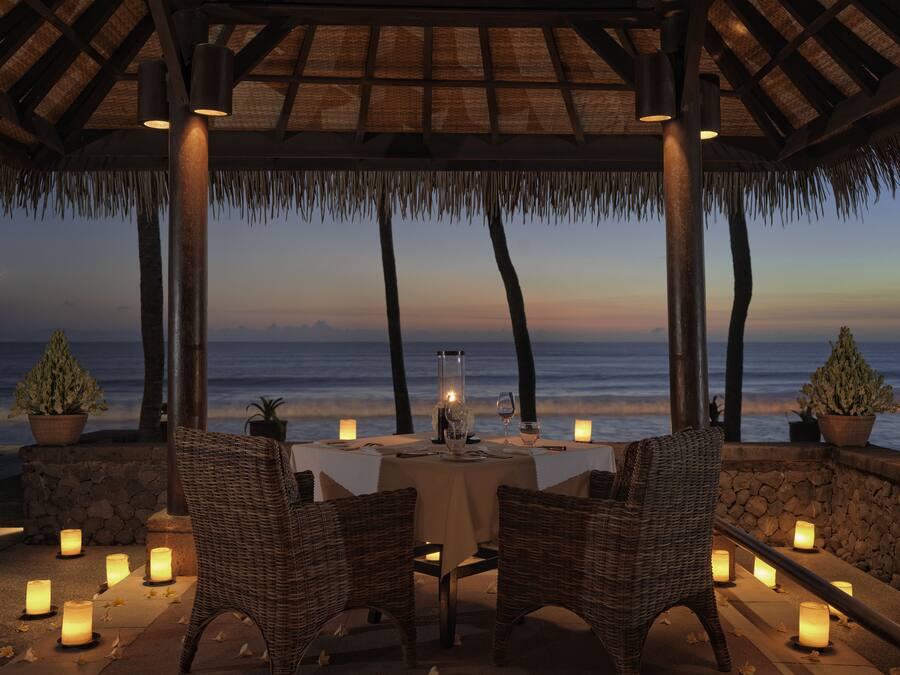 A beachside dining setup with wicker chairs, a table, candles, and a view of the ocean at dusk.