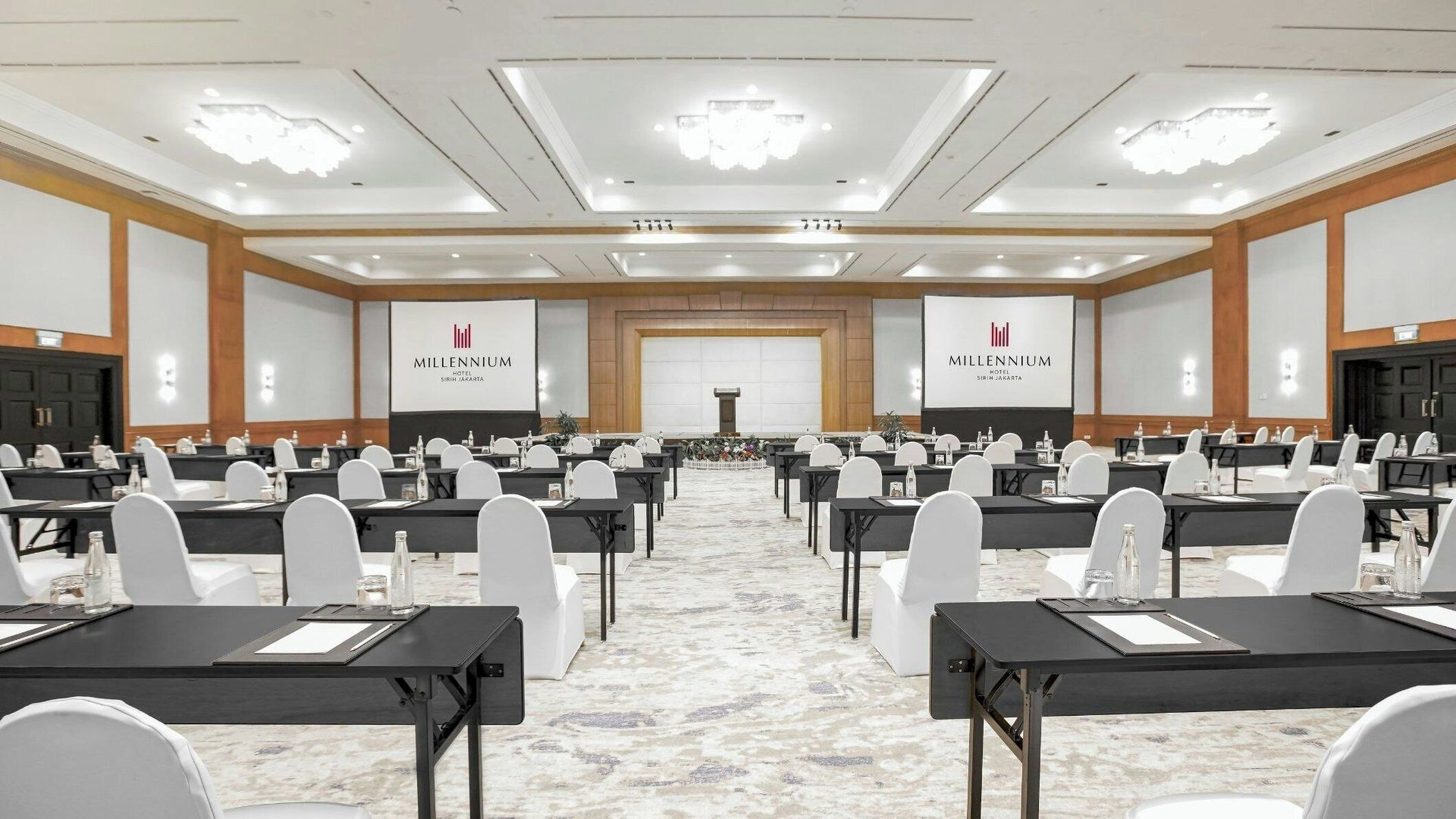 Une salle de conférence équipée de plusieurs tables et chaises, avec une scène à l’avant et un grand écran affichant le nom de l’hôtel.