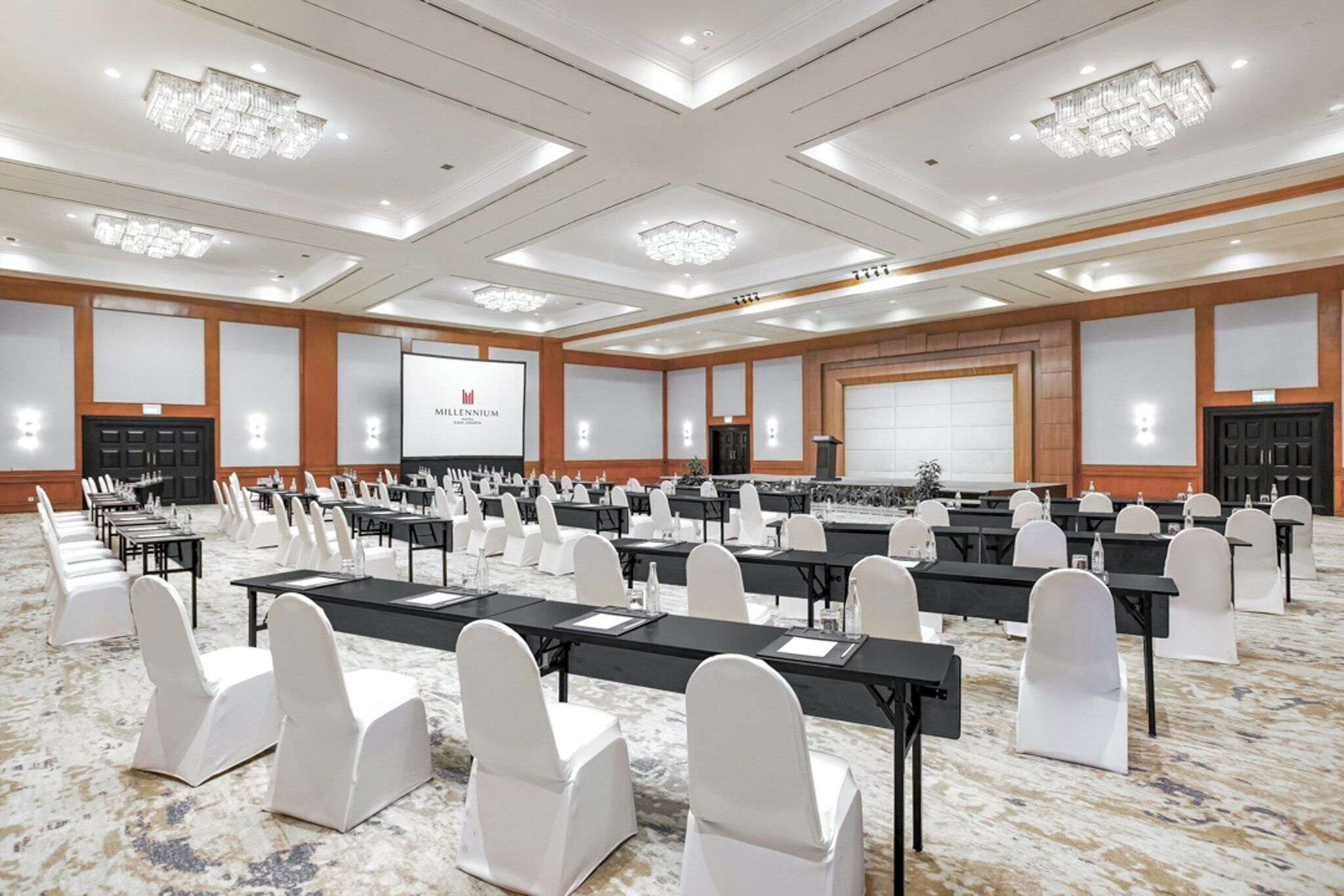 Une grande salle de conférence avec de nombreuses rangées de tables et de chaises, un écran de présentation et des lustres.