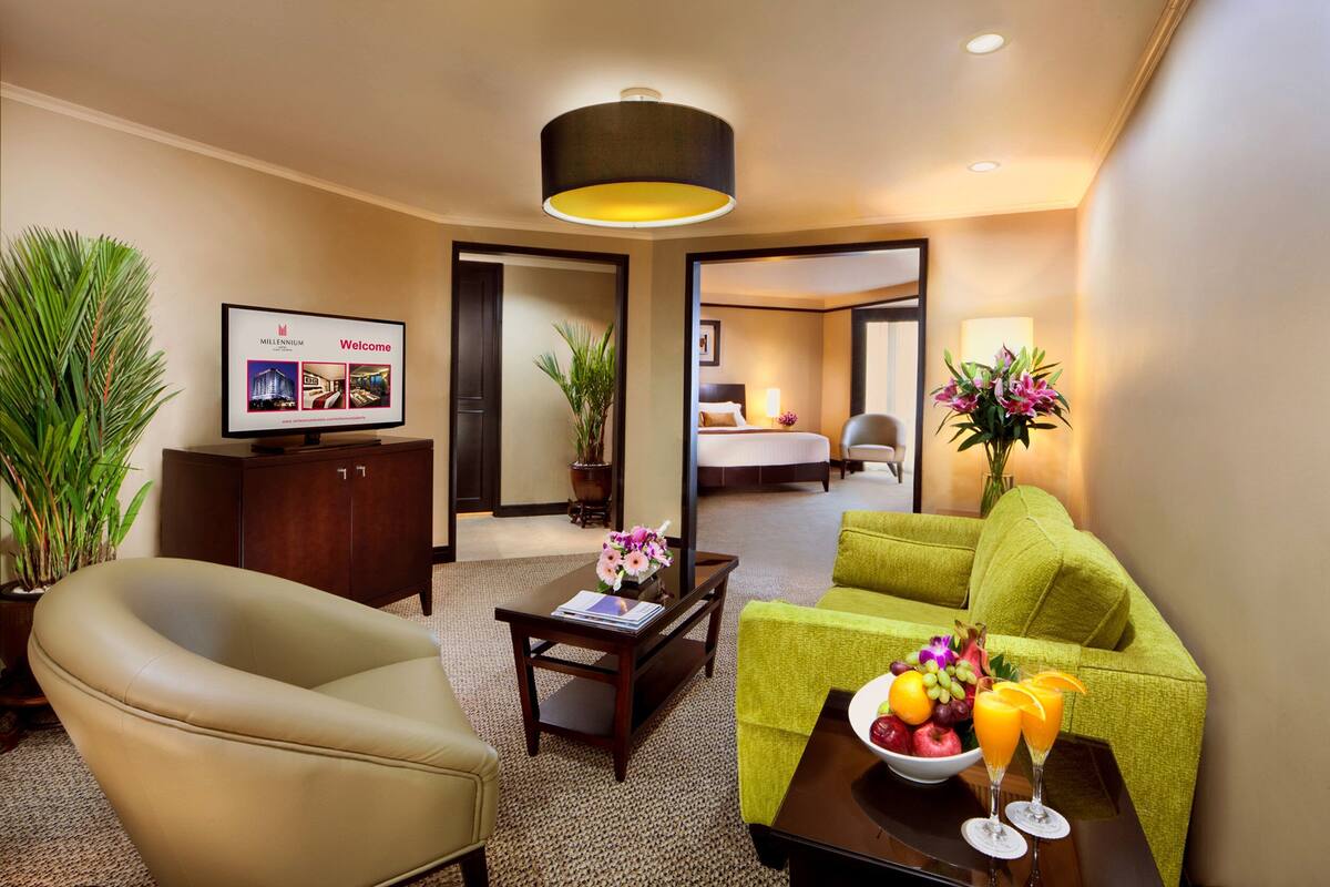 Habitación de hotel con una cama, un escritorio con computadora, un sofá verde, una mesa de centro con fruta y jugo, y un televisor mostrando el mensaje de bienvenida del hotel.
