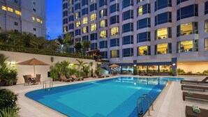 Una piscina con sillas de descanso y sombrillas, rodeada por un edificio de varios pisos y ventanas.