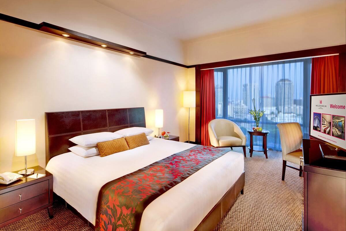Habitación de hotel con una cama grande, un escritorio, una silla y vistas a un paisaje urbano a través de amplias ventanas.