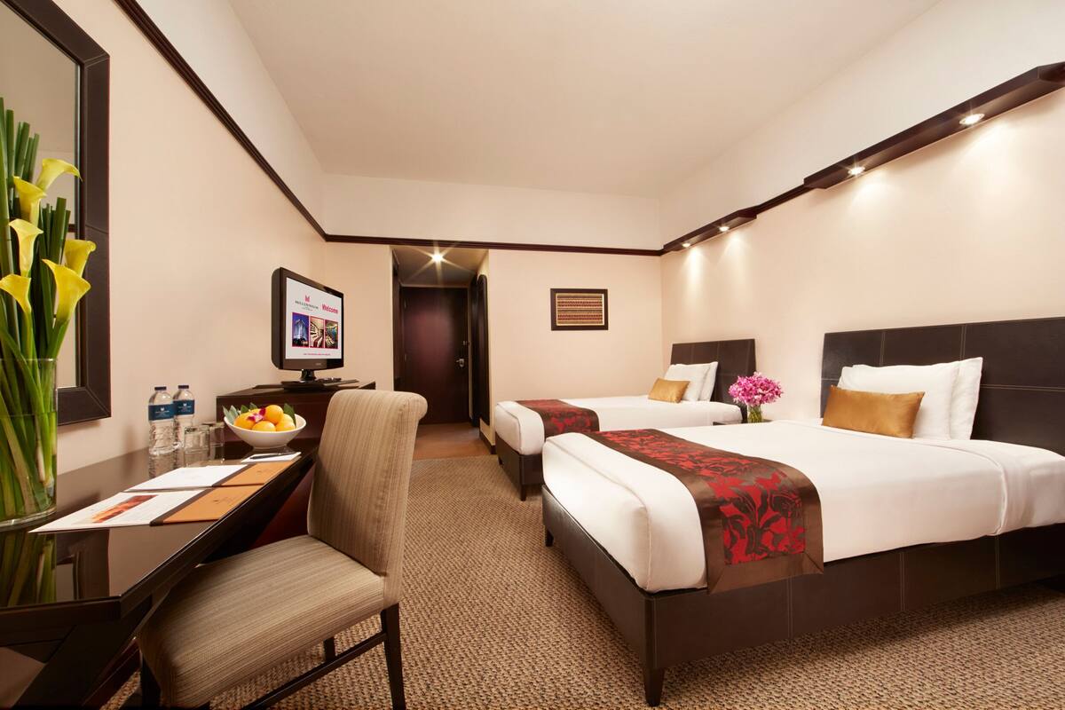 Habitación de hotel con escritorio, dos camas, una silla, un espejo y un jarrón con flores.