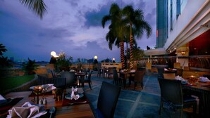 Un área de comedor al aire libre con mesas y sillas, palmeras y vista al horizonte de la ciudad.