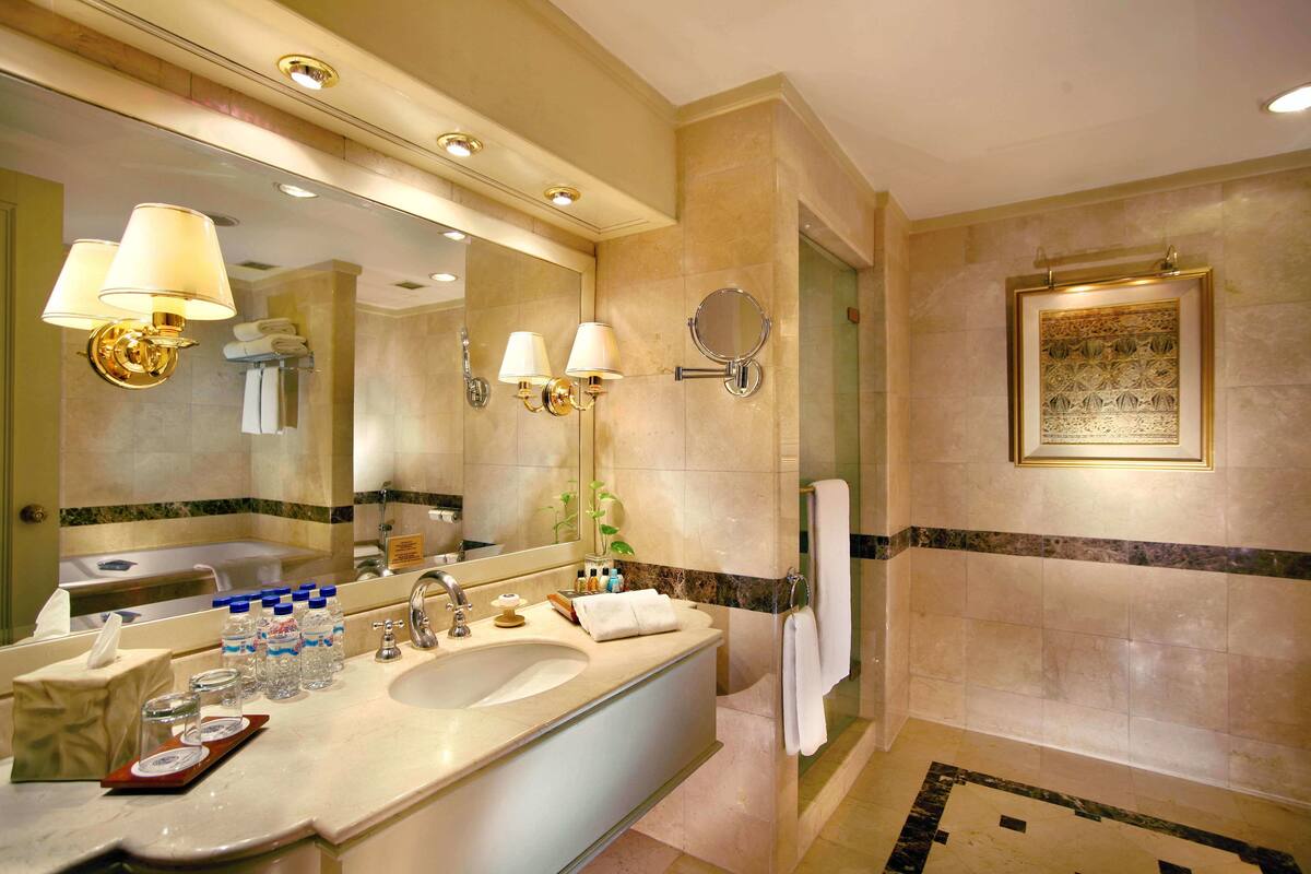 A hotel bathroom with a large mirror, multiple light fixtures, a sink, and a shower area.