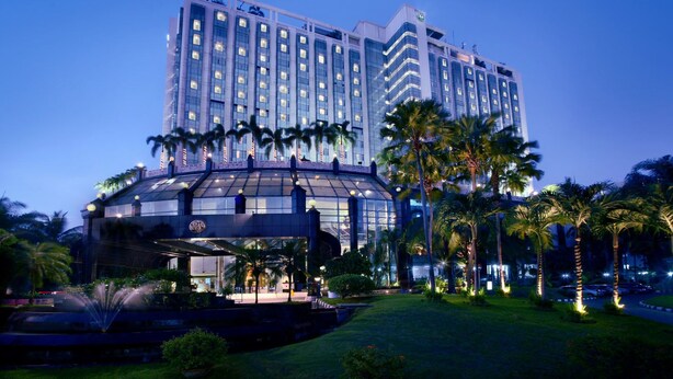 Un hotel moderno con fachada de vidrio, palmeras y una fuente al frente.