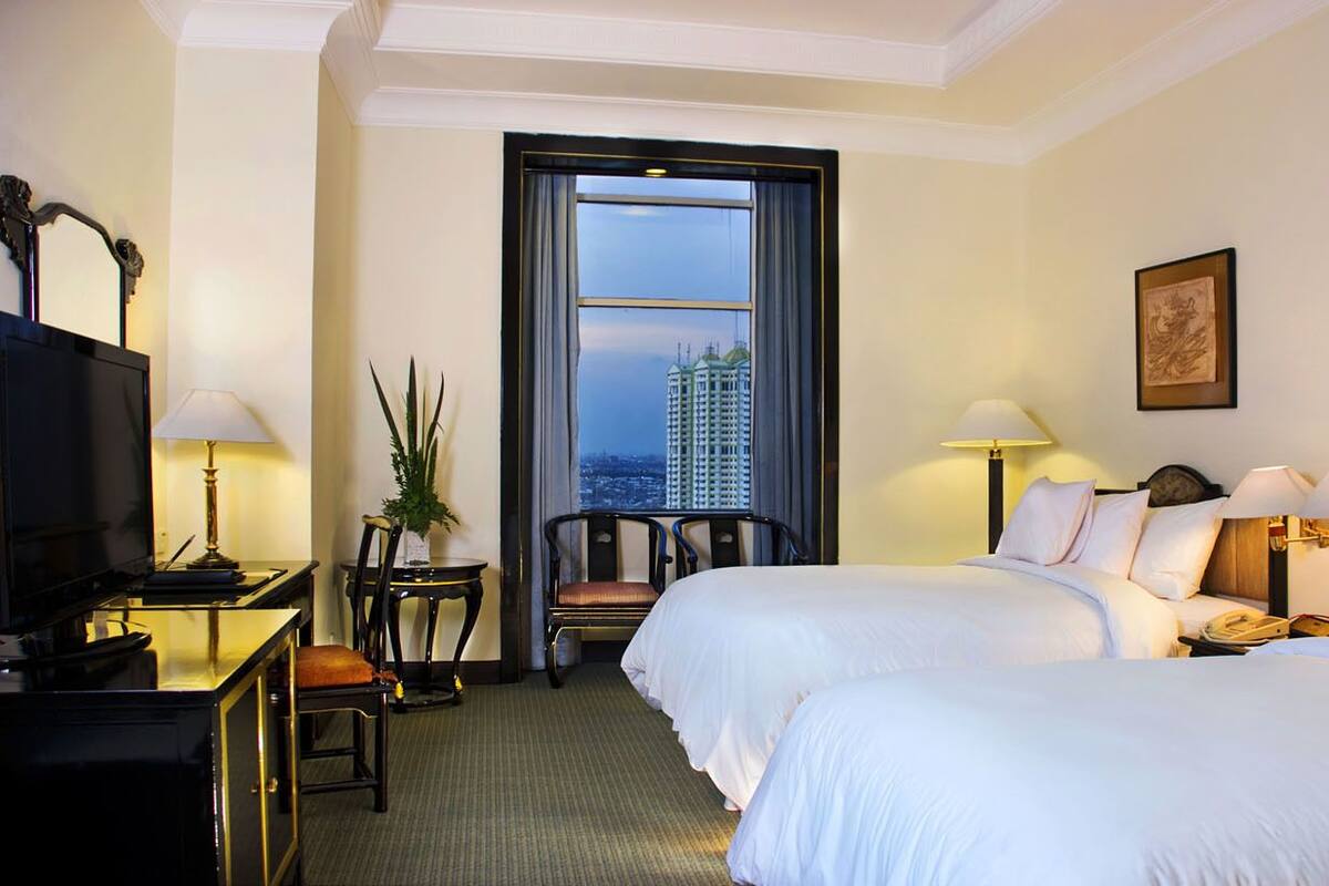 A hotel room with two beds, a desk with a lamp, a chair, a television, and a large window with a city view.
