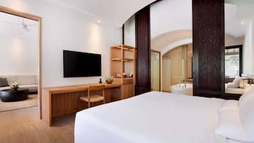 A modern hotel room with a large bed, a flat-screen TV, a desk with a chair, a potted plant, and a shower area.