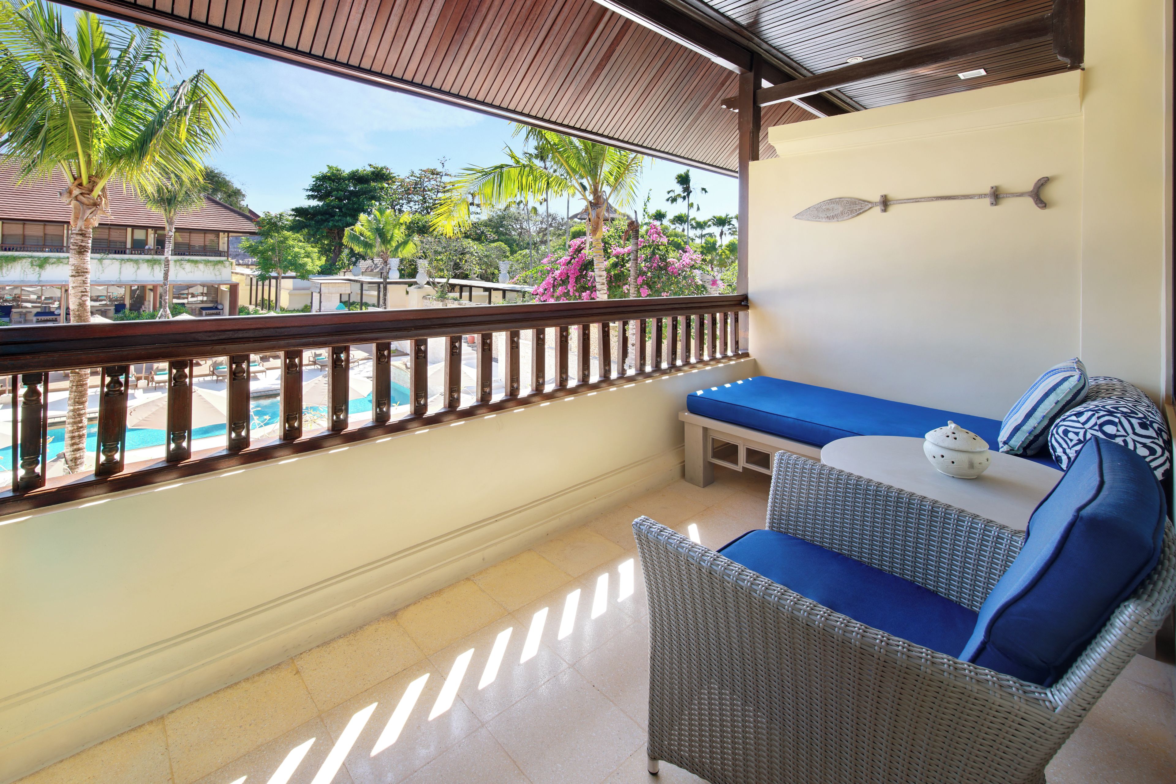 A balcony with a wicker sofa, a small table, and a view of a pool and tropical vegetation.