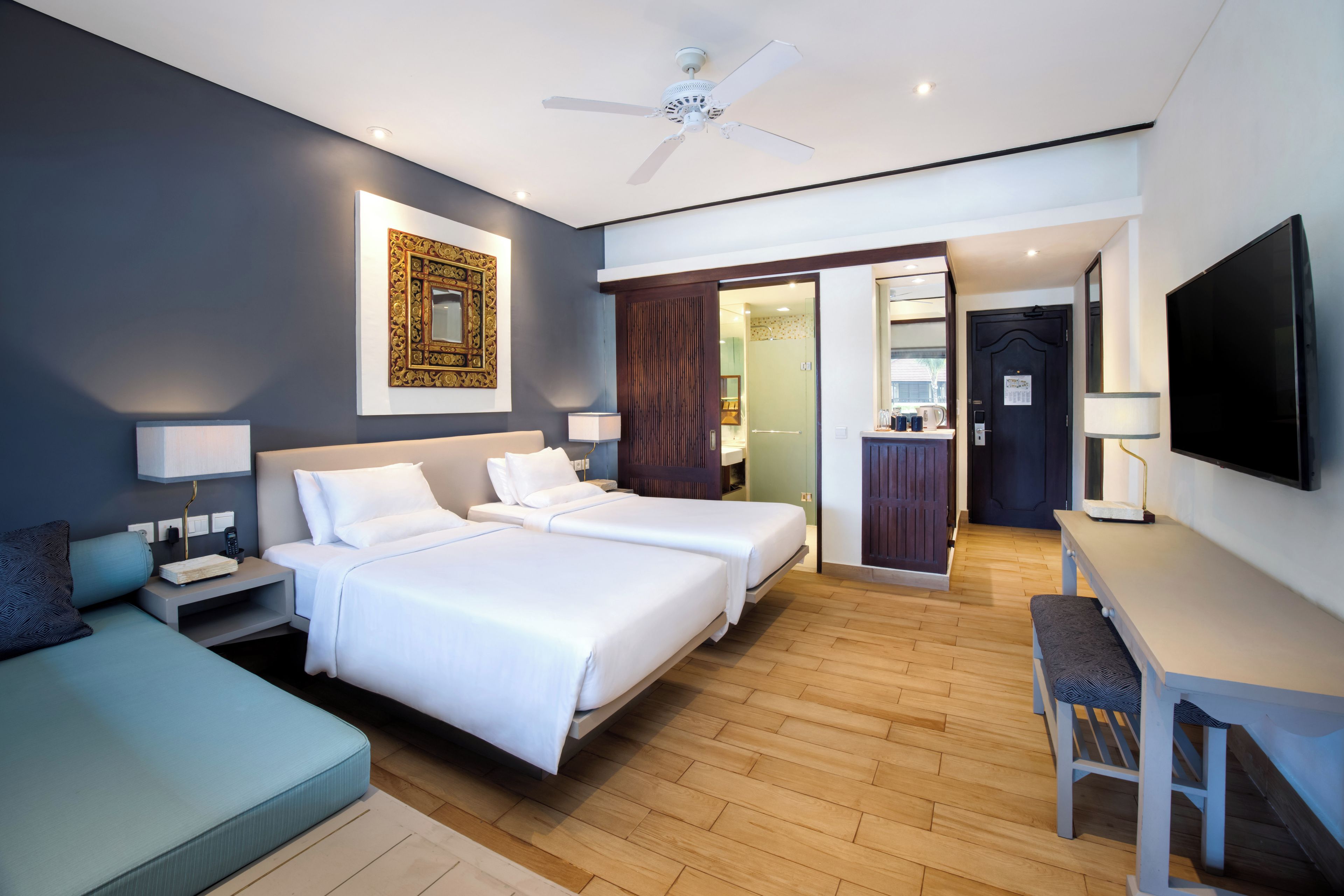 A hotel room with two beds, a desk, a TV, a ceiling fan, and a bathroom visible through an open door.