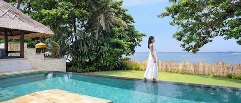 A pool area with a clear view of the sea, surrounded by lush greenery and a thatched-roof structure.