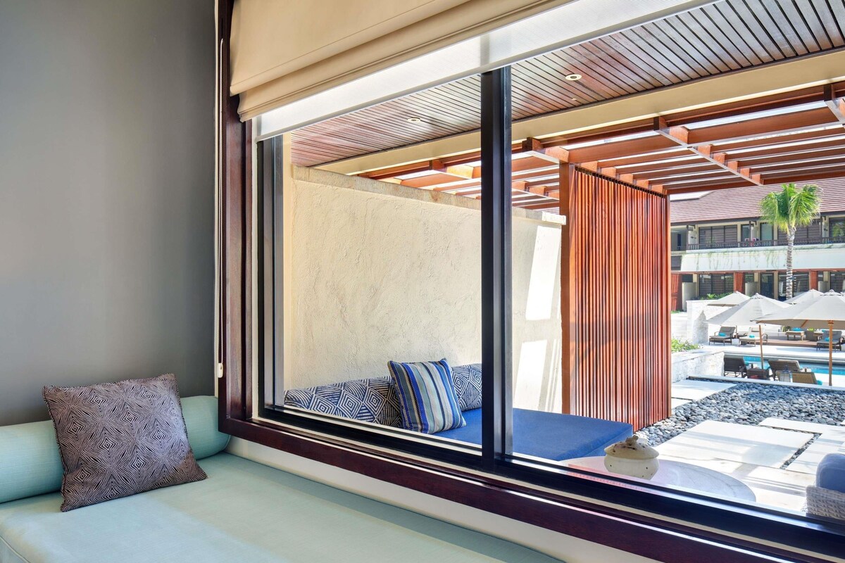 A modern hotel room with a large window, a sofa with patterned cushions, and a view of the outdoor pool area.