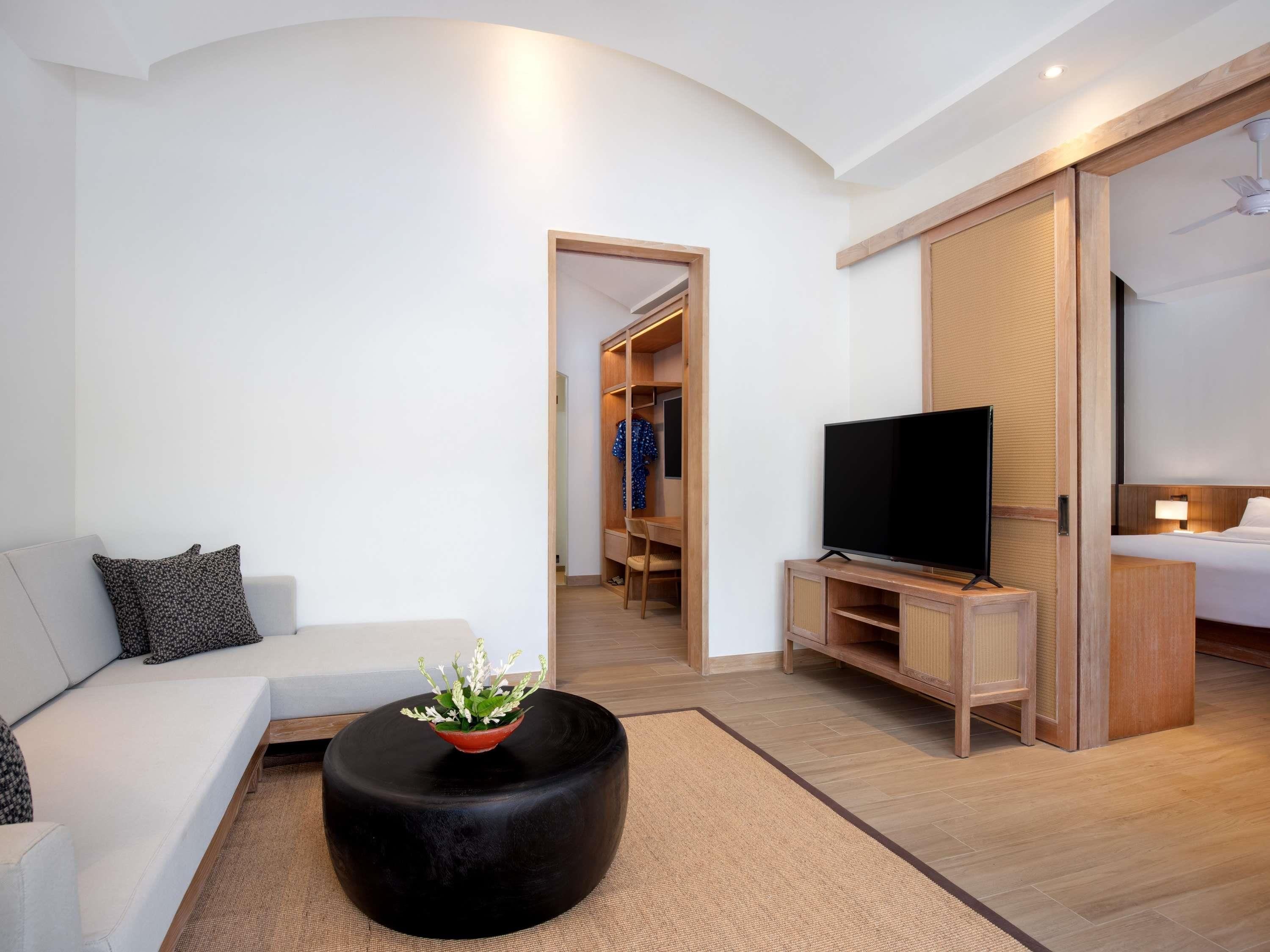 Een moderne hotelkamer met een bank, een zwart voetenbankje met een plant, een televisie, een houten garderobe en een bed.