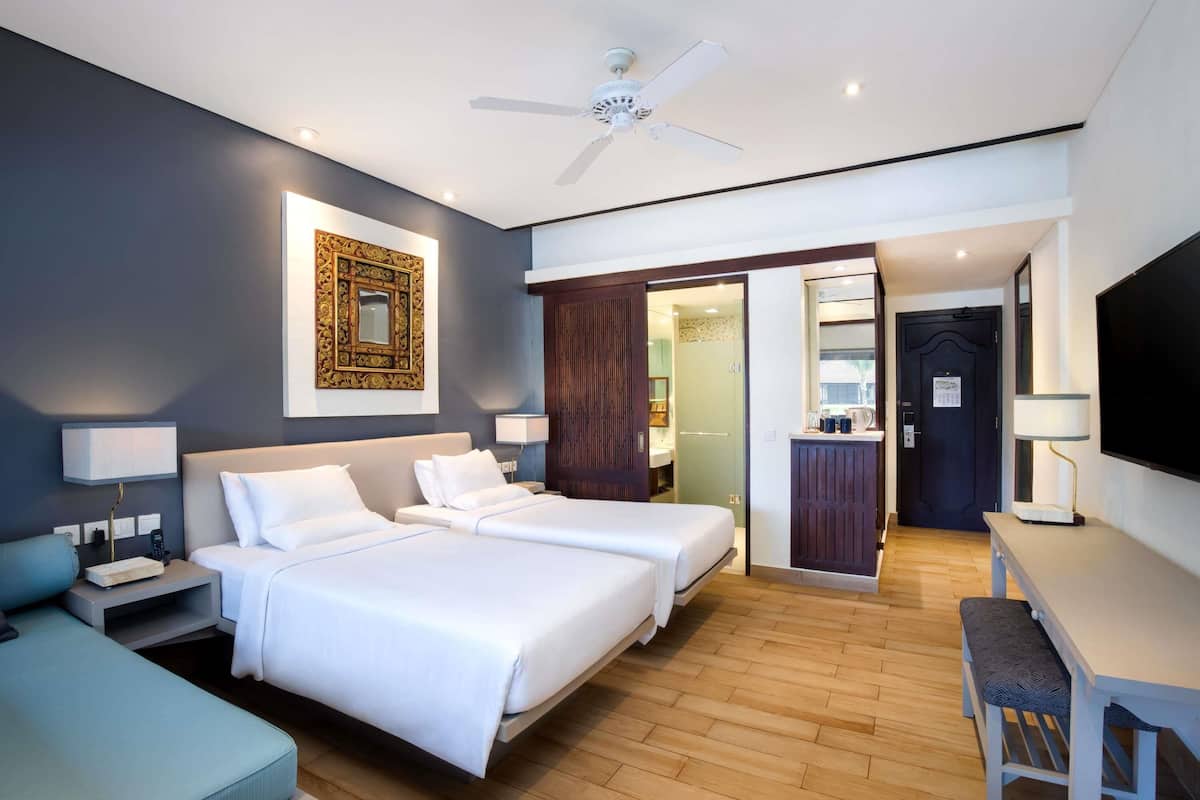 Habitación de hotel moderna con dos camas, un escritorio, una silla, un televisor, un ventilador de techo y un baño visible a través de una puerta abierta.
