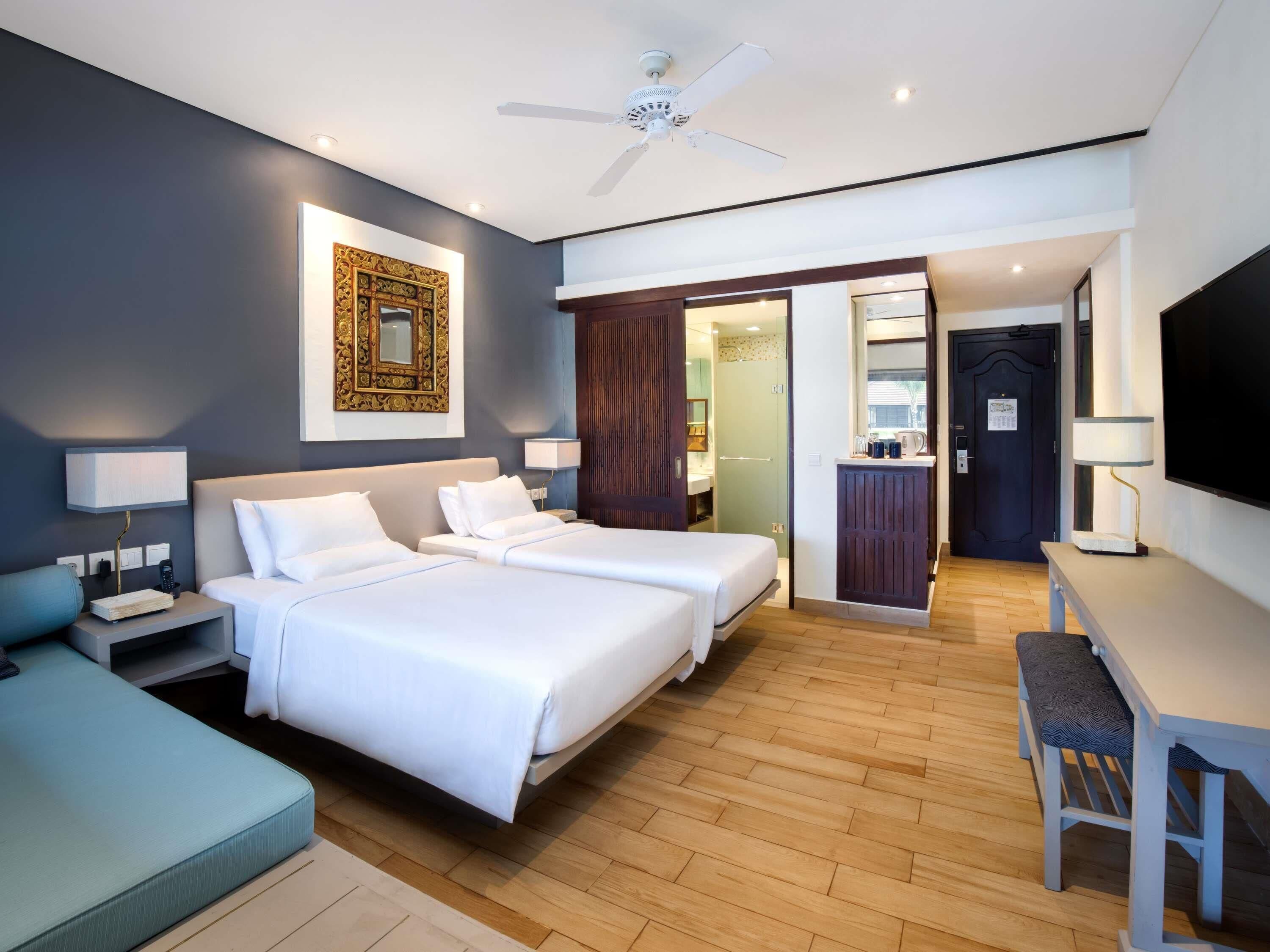 Une chambre d’hôtel avec deux lits, un bureau, une chaise, une télévision, un ventilateur de plafond et une salle de bain visible à travers une porte ouverte.