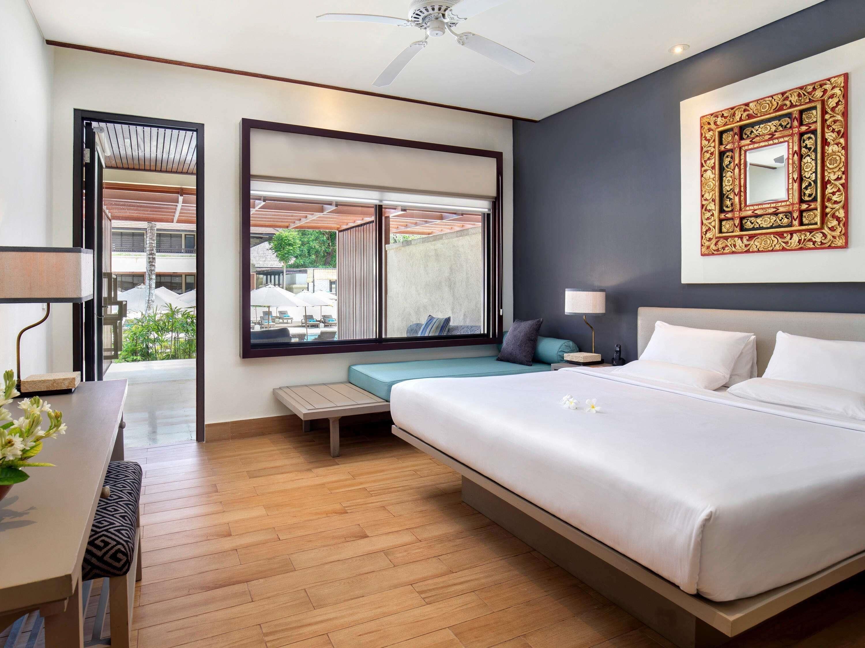 Une chambre d’hôtel moderne dotée d’un grand lit, d’un bureau avec une lampe, d’une fenêtre donnant sur l’extérieur et d’un ventilateur de plafond.