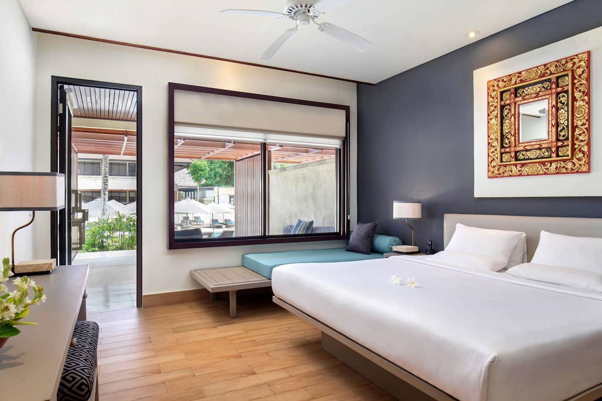 Une chambre d’hôtel moderne dotée d’un grand lit, d’un bureau avec une lampe, d’une fenêtre donnant sur l’extérieur et d’un ventilateur de plafond.