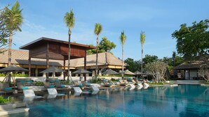A resort pool area with lounge chairs and umbrellas, surrounded by palm trees and a building with a thatched roof. - Novotel Bali Benoa (Nusa Dua)