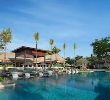 A resort pool area with lounge chairs and umbrellas, surrounded by palm trees and a building with a thatched roof.