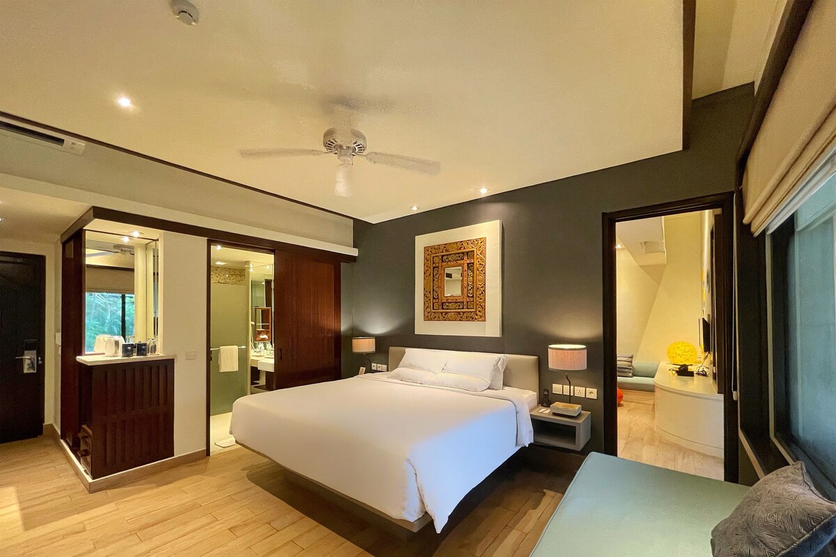 A modern hotel room with a large bed, bedside tables, a ceiling fan, and a bathroom visible through an open door.