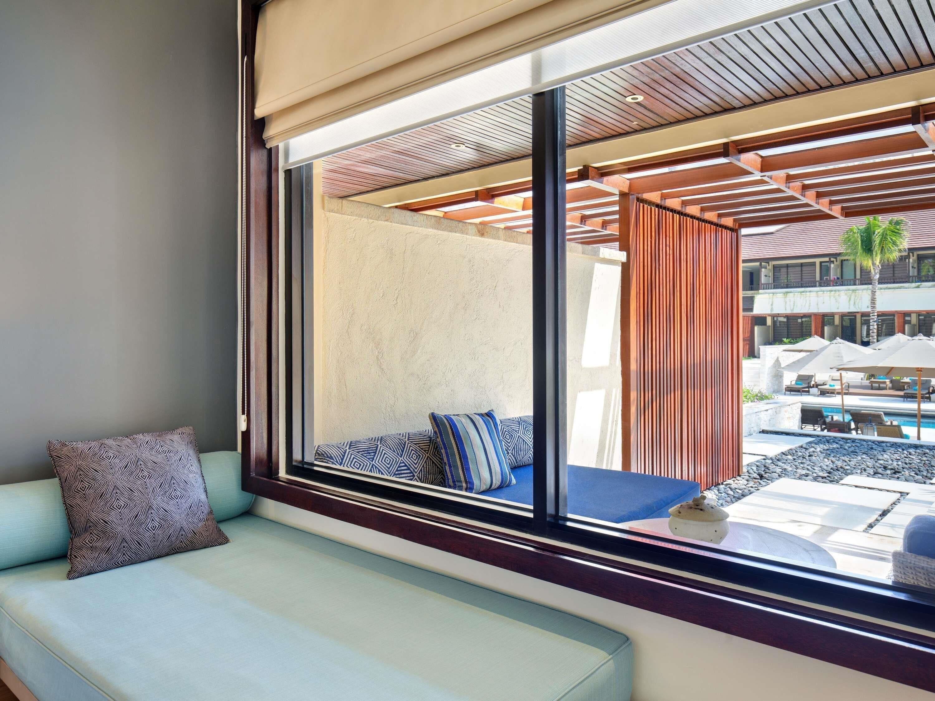 A modern hotel room with a large window, a sofa with patterned cushions, and a view of the outdoor pool area.