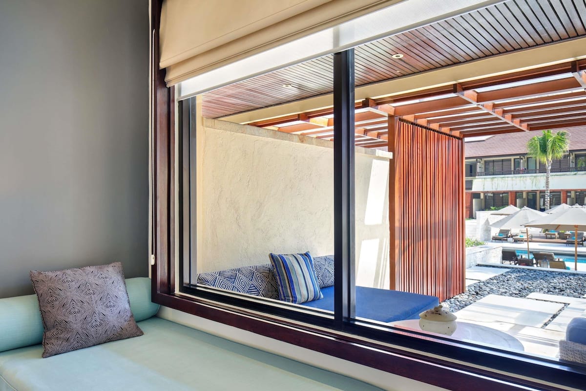 A modern hotel room with a large window, a sofa with patterned cushions, and a view of the outdoor pool area.