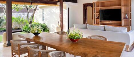 A dining area with a wooden table and wicker chairs, a sofa, and a TV mounted on a wooden cabinet.