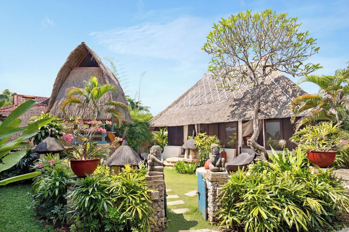 A tropical garden with thatched-roof structures, lush greenery, and stone statues.
