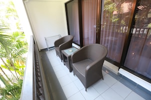 A balcony with wicker furniture, a glass table, and a view of palm trees and buildings.