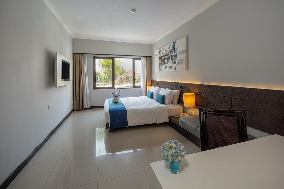 A hotel room with a bed, bedside table, lamp, and a view of the outdoors.