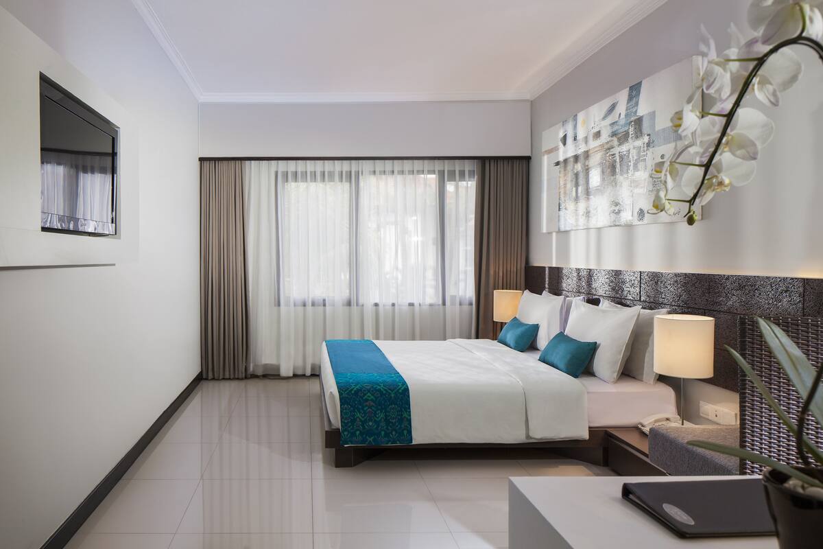 A modern hotel room with a large bed, a desk with a laptop, a nightstand, and a potted plant.