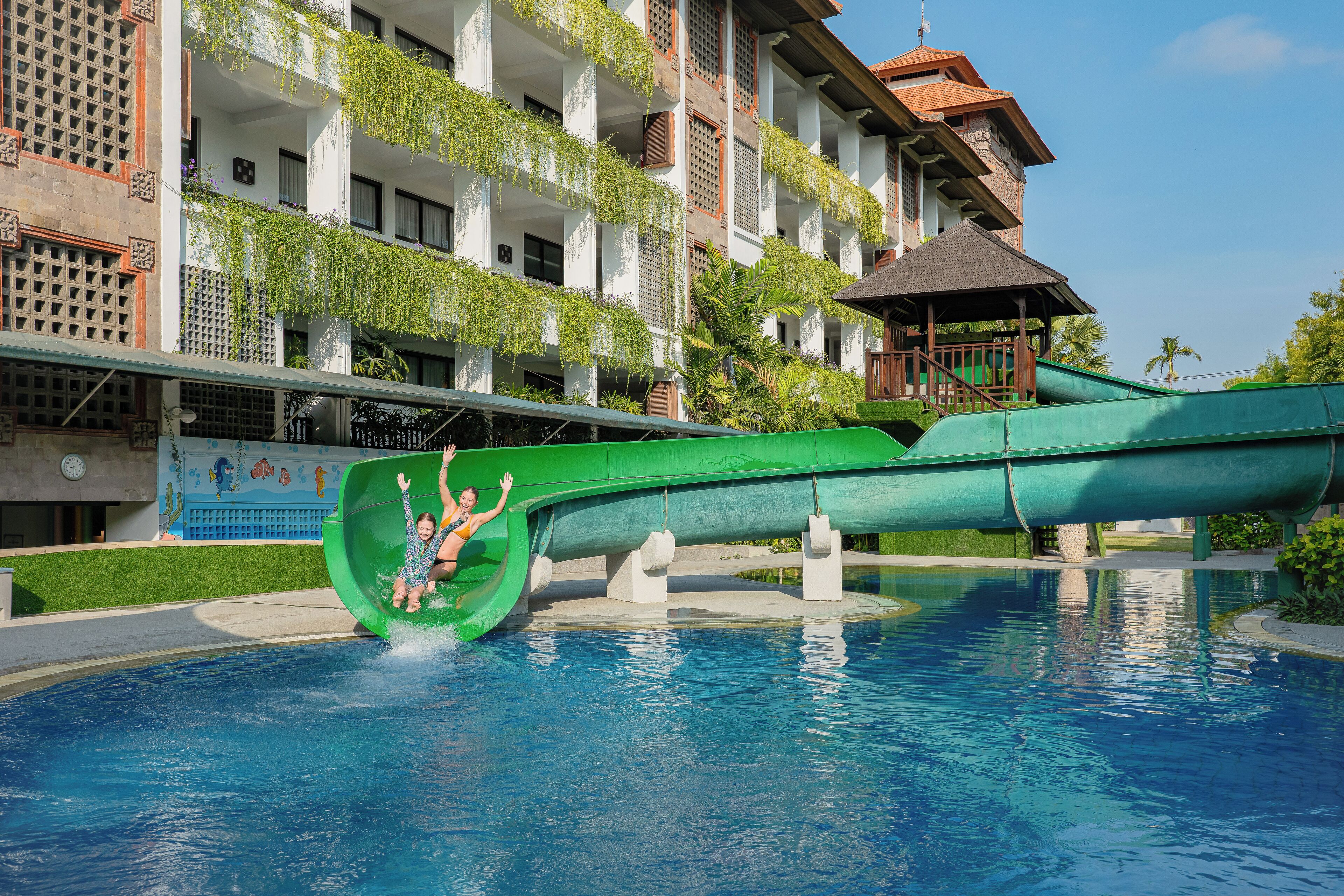 A swimming pool with a green water slide and a building with balconies in the background.