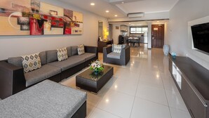 A modern living room with a sofa set, a coffee table with flowers, a TV, and a kitchen area in the background.