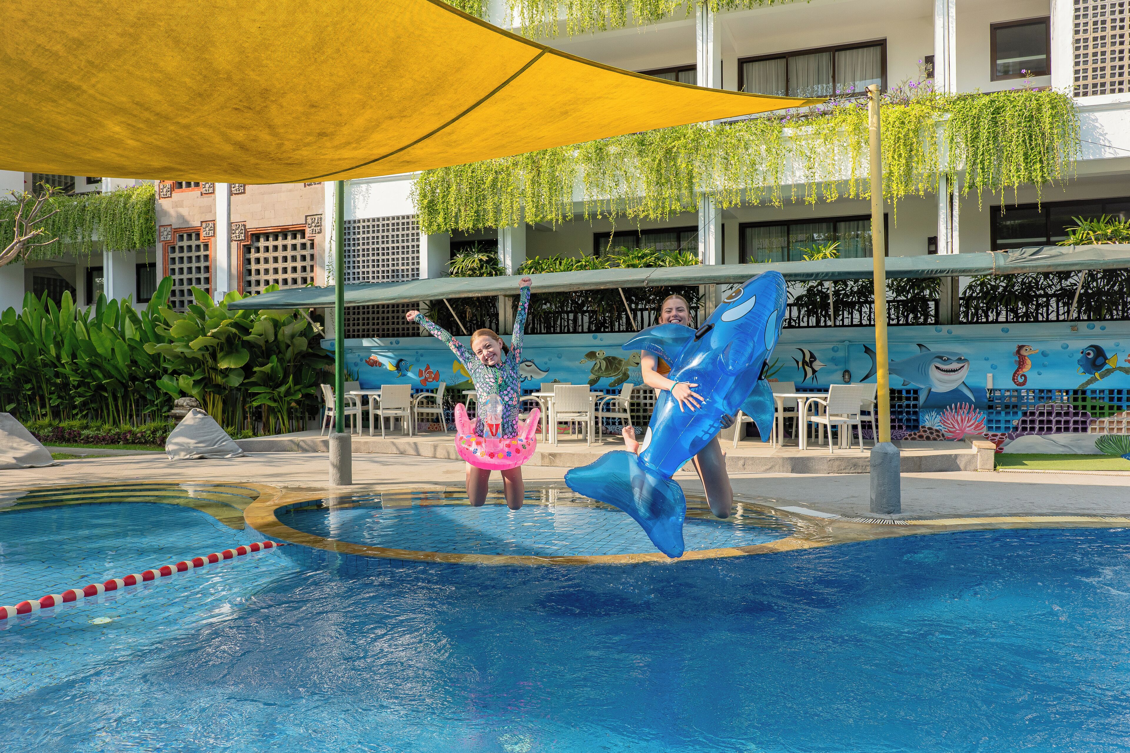 Zona de piscina con plataforma de salto, una tolda amarilla y un mural con vida marina.