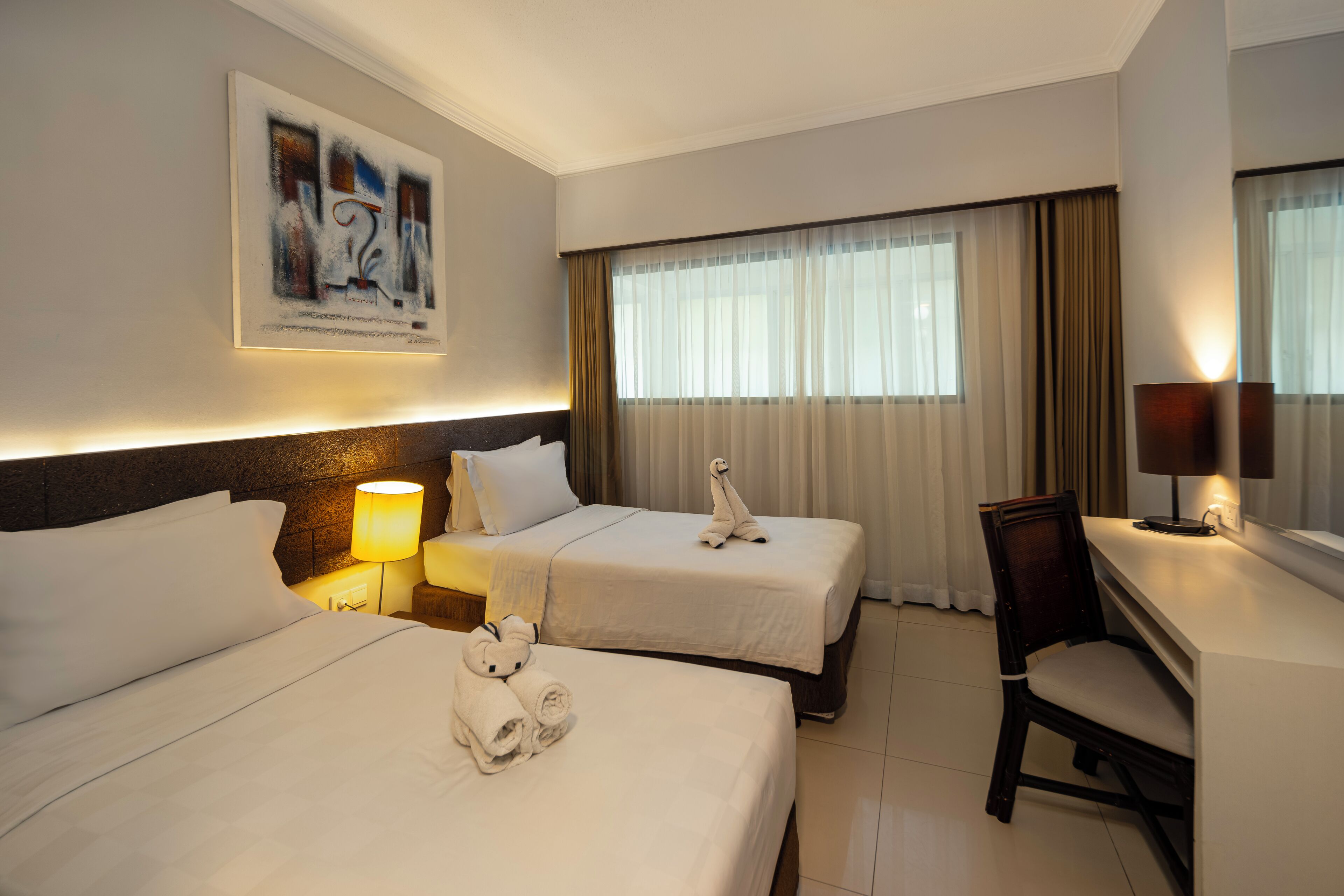 A hotel room with two beds, a desk, a chair, a lamp, and a painting on the wall.